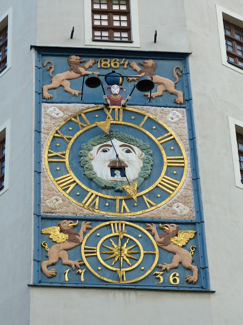 043.jpg - 043 Barokowy zegar z 1693 r. Powstał na okoliczność uczczenia zwycięstwa Szwedów w wojnie z Brandenburgią. Zielony maszkaron porusza oczami zgodnie z ruchem dużej wskazówki godzinowej. W ustach ma cyfrę pokazującą dzień miesiąca. Znajdujący się powyżej błazen wybija lewą ręką godziny, a prawą kwadranse. Ozdobiona gryfami dolna tarcza nie ma skali. Dokładniej pokazuje kwadranse niż minuty. Daty 1736 i 1864 r. dotyczą remontów zegara